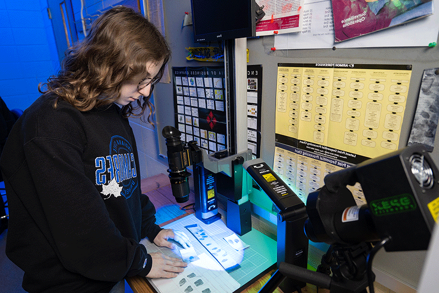 A female student with dark brown hair and glasses, and wearing a black Sycamores sweatshirt, stands in a dim room analyzing fingerprints beneath lights on her desk. On the wall in front of her are assorted charts with graphical information and images.