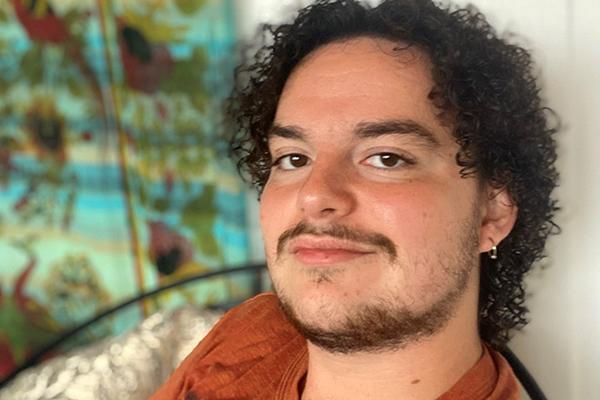 Close-up of a student with curly brown hair with a matching beard and moustache. The student is staring at the camera and wears an orange shirt. In the background is a blue, red, and yellow floral art piece.